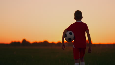 Boy-football-player-with-the-ball-goes-across-the-field-the-sunset-camera-steadicam-follows-the-boy.-Boy-at-dawn-watching-the-sunrise-standing-with-a-soccer-ball.